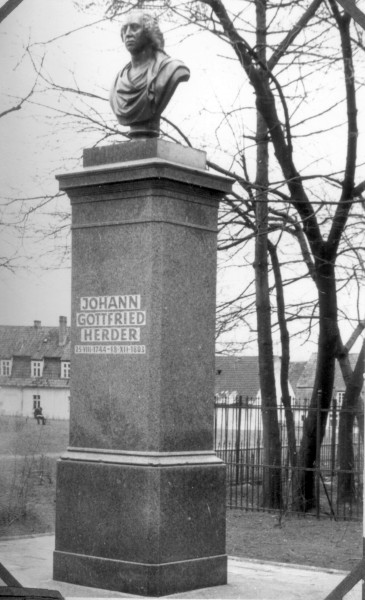 Popiersie Johanna Gottfrieda Herdera znajduje się na wysokim kamiennym cokole w parku, w tle widać bezlistne drzewa i budynki.
