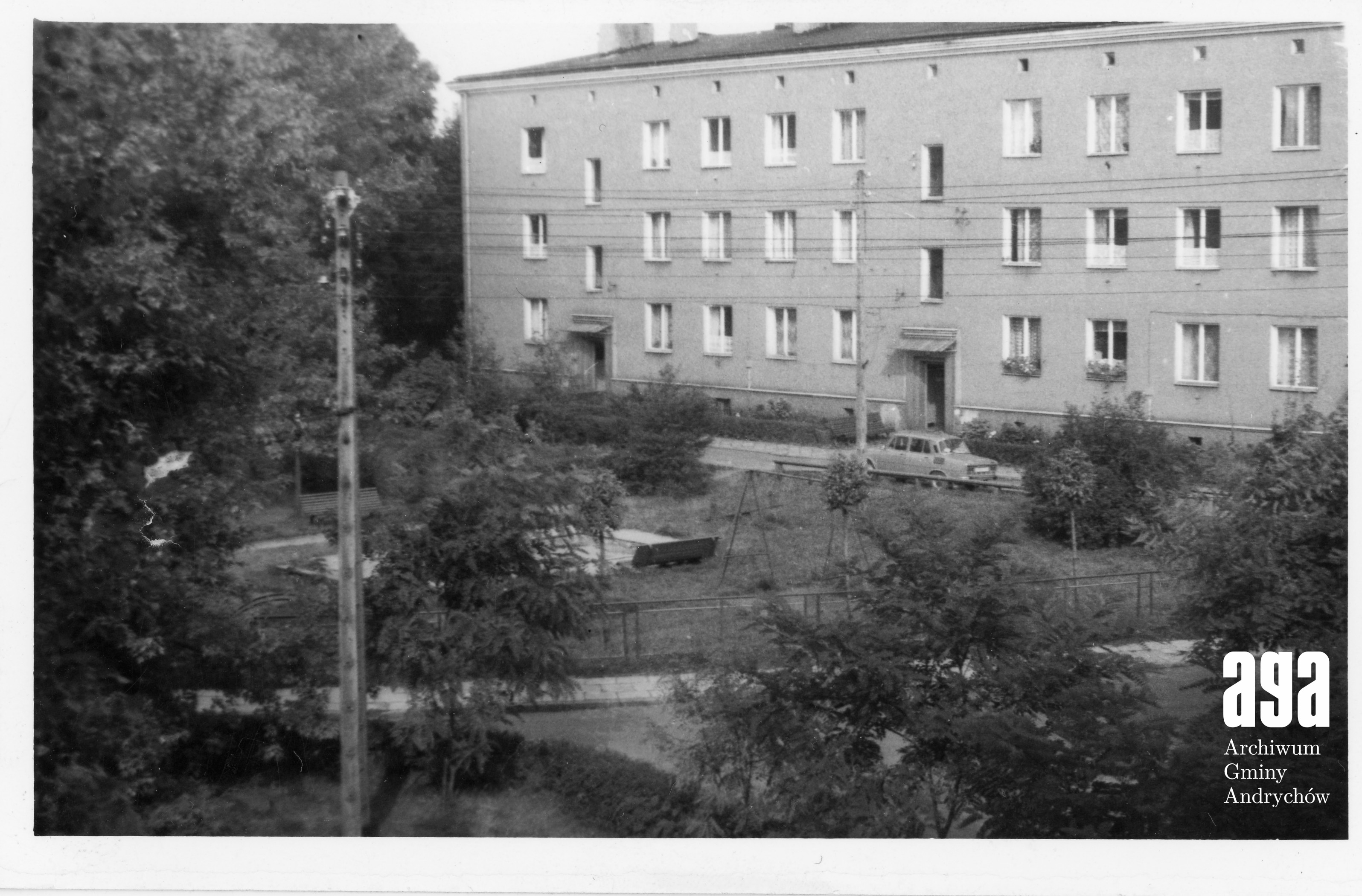 Czarno-białe zdjęcie trzypiętrowego ceglanego budynku mieszkalnego z drzewami i ogrodem. Przed budynkiem zaparkowany jest pojedynczy samochód.