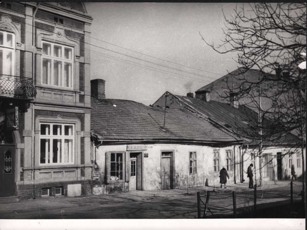 Czarno-białe zdjęcie starej ulicy z rzędem zniszczonych budynków, dwiema osobami idącymi i nagimi gałęziami drzew po prawej stronie.