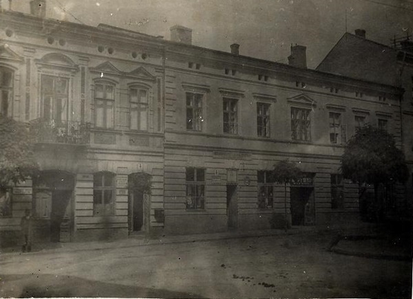 Sepiowe zdjęcie przedstawiające stary, dwupiętrowy budynek z kilkoma oknami i drzwiami, drzewami z przodu i zachmurzonym niebem.