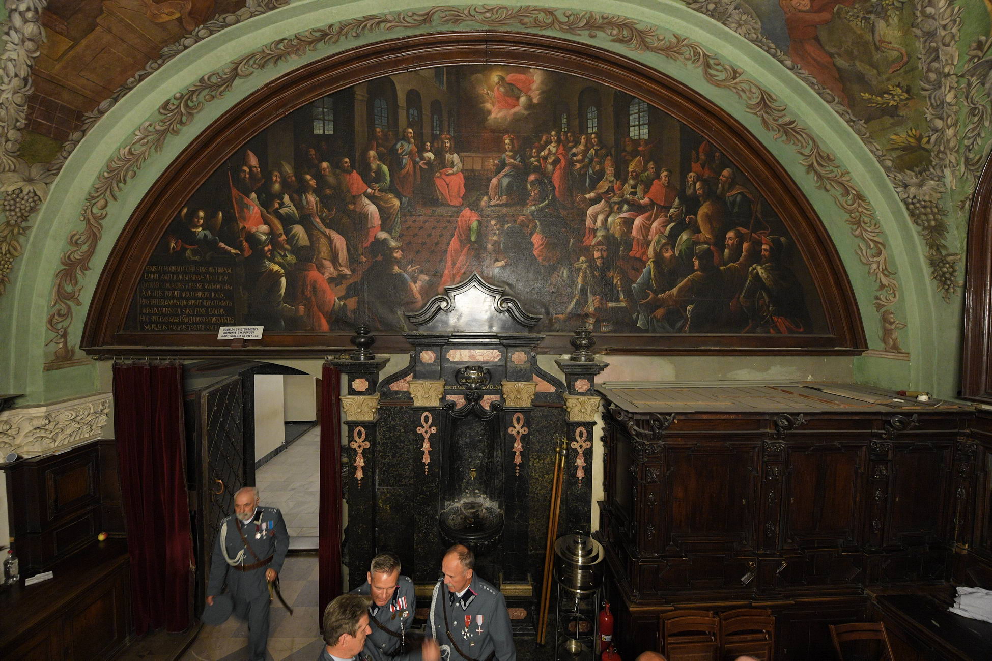 Historyczne pomieszczenie z dużym łukowatym obrazem przedstawiającym zgromadzenie ludzi. Kilka osób w mundurach rozmawia poniżej. Wystrój obejmuje ozdobną stolarkę i klasyczne dzieła sztuki.