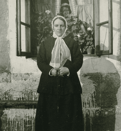 Kobieta w tradycyjnym stroju stoi na zewnątrz budynku przed oknem. W oknie widać rośliny oraz ukrytą twarz kobiety. Link do podstrony archiwum Biblioteki w Pyzdrach