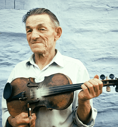 Starszy mężczyzna, widoczny od pasa w górę, pozuje ze skrzypcami. Trzyma instrument poziomo na wysokości piersi. Stoi na tle bielonej wiejskiej chaty. Link do wyników wyszukiwania z tym hasłem
