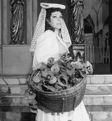 Ubrana na biało aktorka stoi na schodach scenografii i trzyma w rękach kosz wypełniony papierowymi kwiatami. Link do podstrony archiwum Fundacji Sztuki, Przygody i Przyjemności ARTS