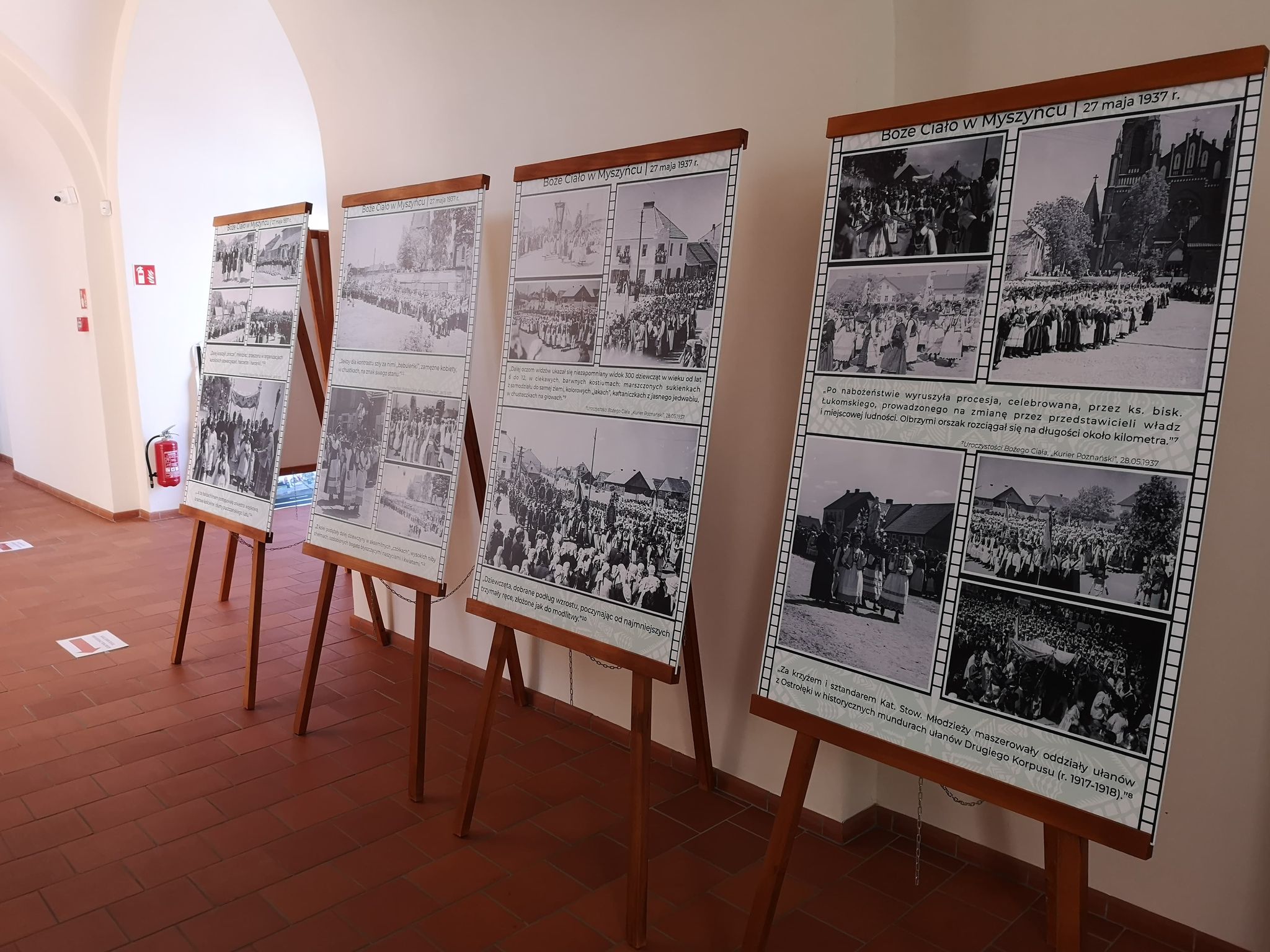 W korytarzu z kafelkową podłogą ustawiono cztery sztalugi, na których umieszczono czarno-białe historyczne fotografie i teksty.