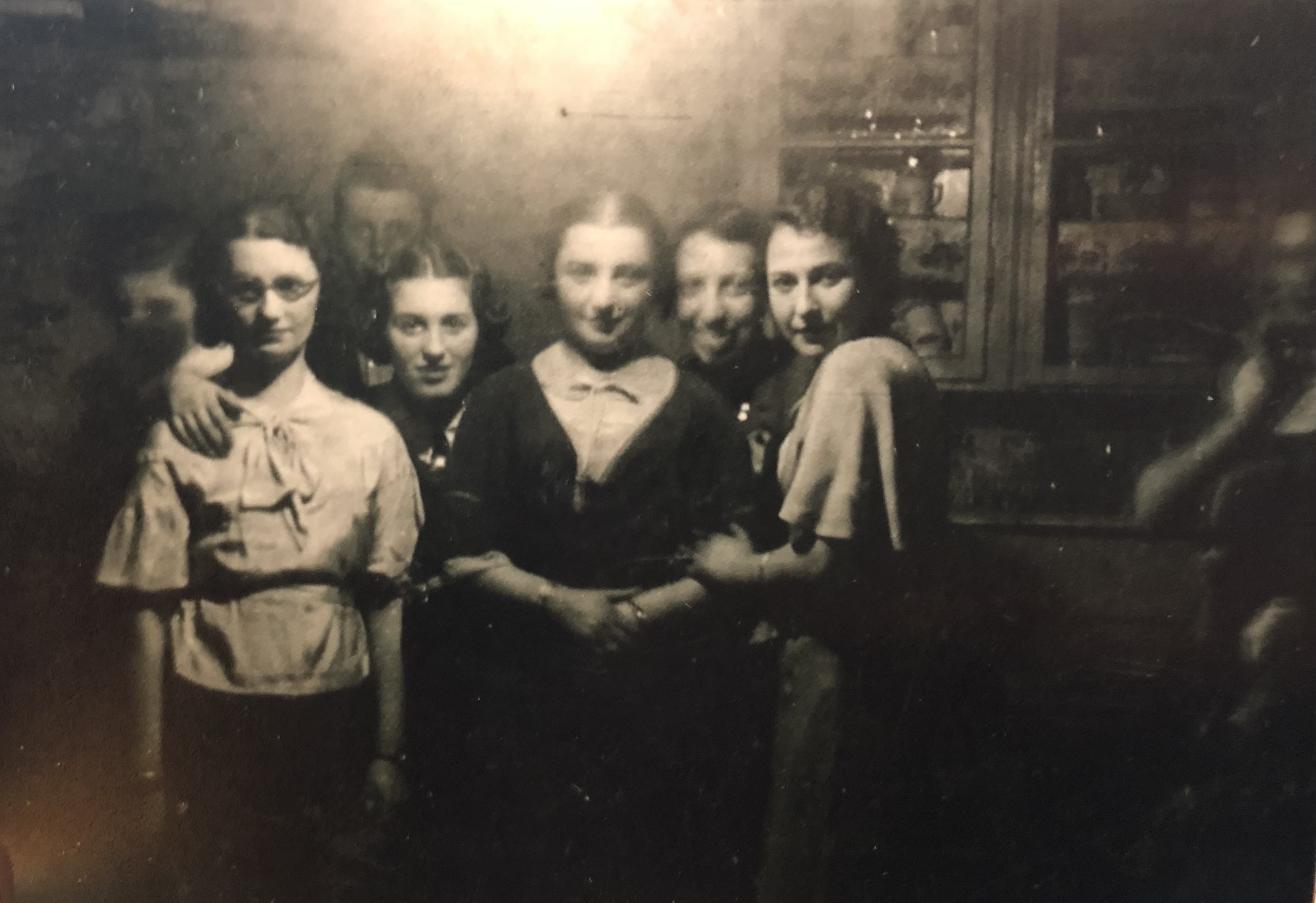 Grupa sześciu młodych kobiet stoi blisko siebie w słabo oświetlonym pokoju, w tle widać szafkę.