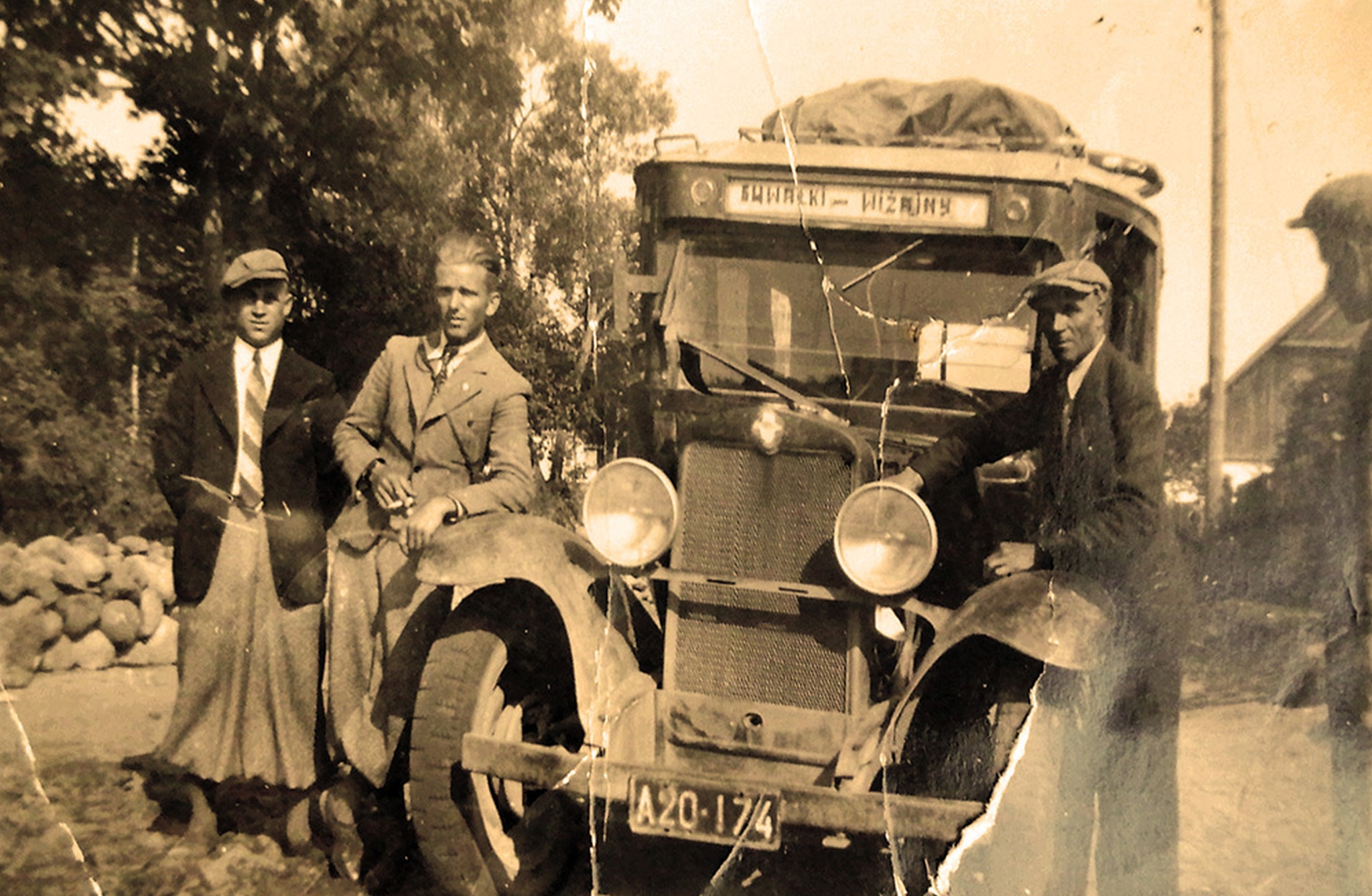 Three men are standing by an old bus with the license plate "A20-174". The bus has luggage on the roof and trees in the background. The photo seems old and sepia.