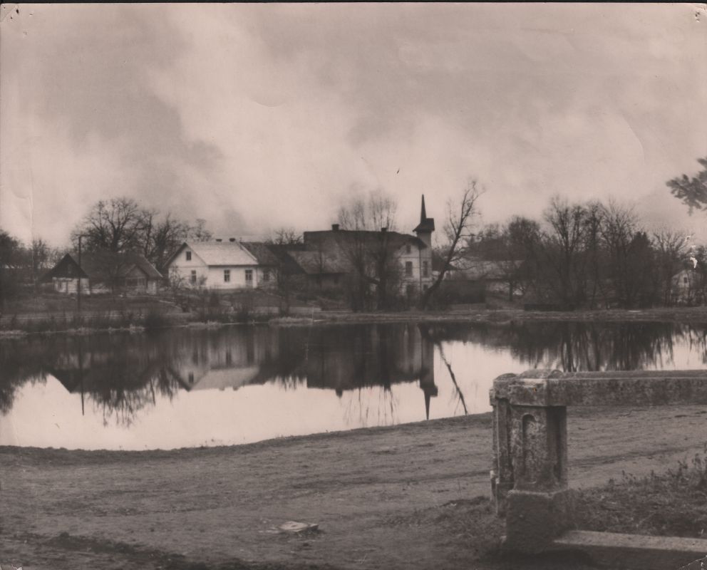 Czarno-białe zdjęcie wioski z domami i wieżą kościelną odbitą w stawie. Nagie drzewa rosną wzdłuż brzegu wody, a na pierwszym planie znajduje się kamienna konstrukcja.